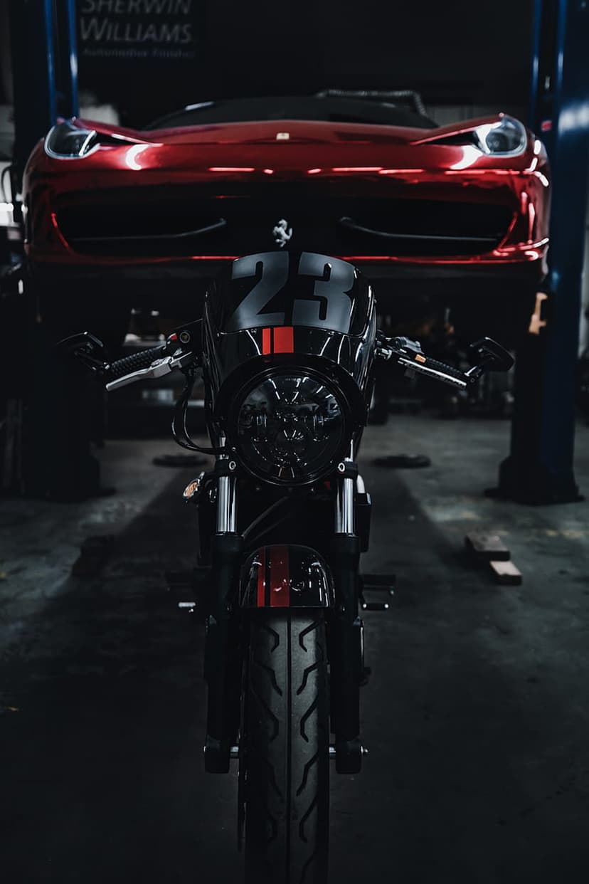 Black racing motorcycle number 23 parked in front of a red Ferrari on a car lift inside a dark workshop.