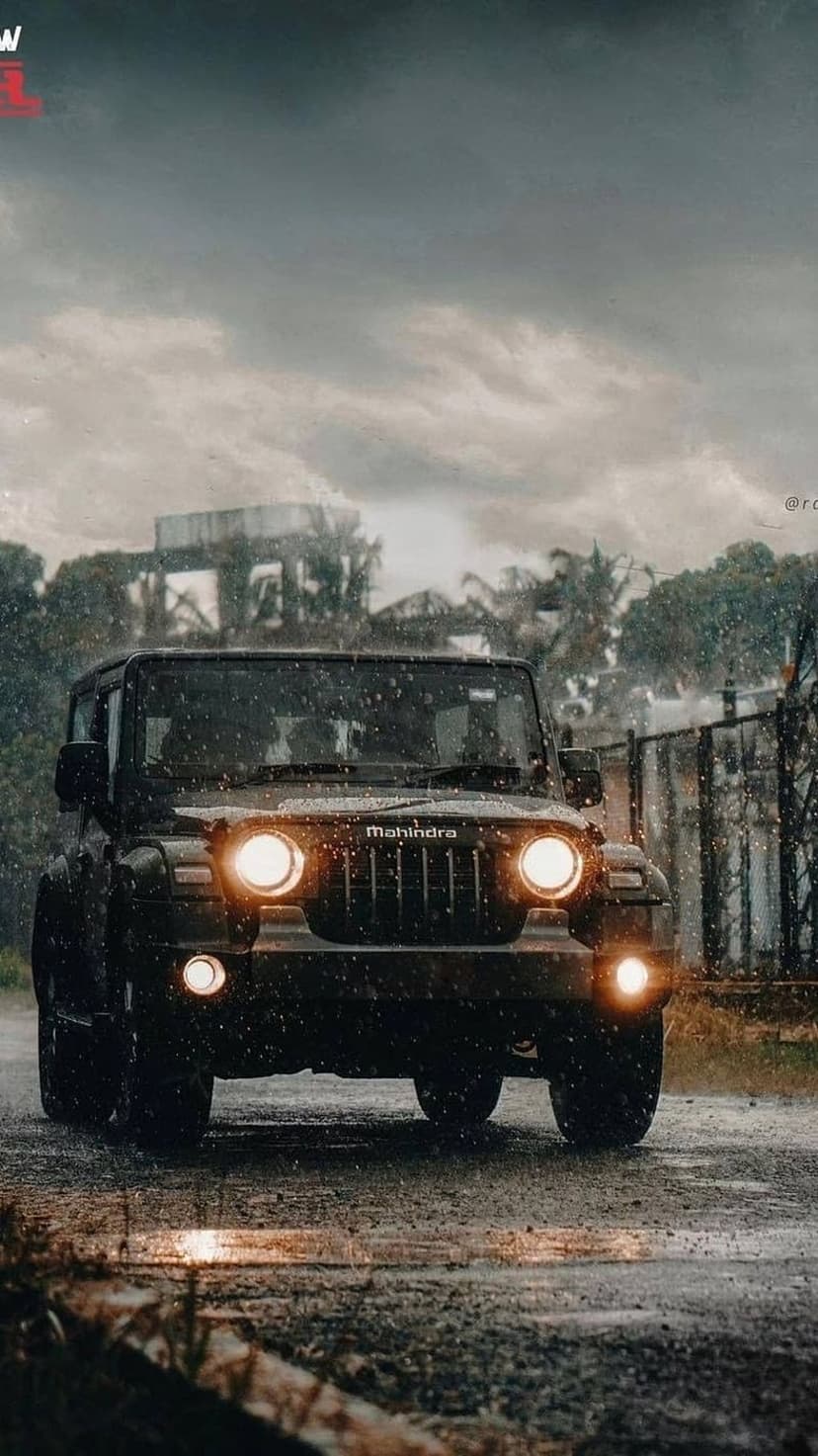 Black Mahindra Thar SUV driving on a wet road during a heavy rainstorm with headlights glowing in the dark fog.
