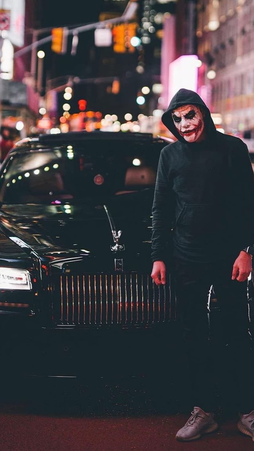 Man in Joker makeup and black hoodie stands by a sleek black sports car against a blurred neon city background.
