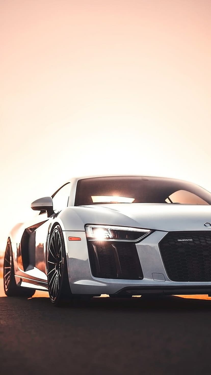 White Audi R8 sports car with glowing headlights on asphalt under a peach sunset sky at a low camera angle.