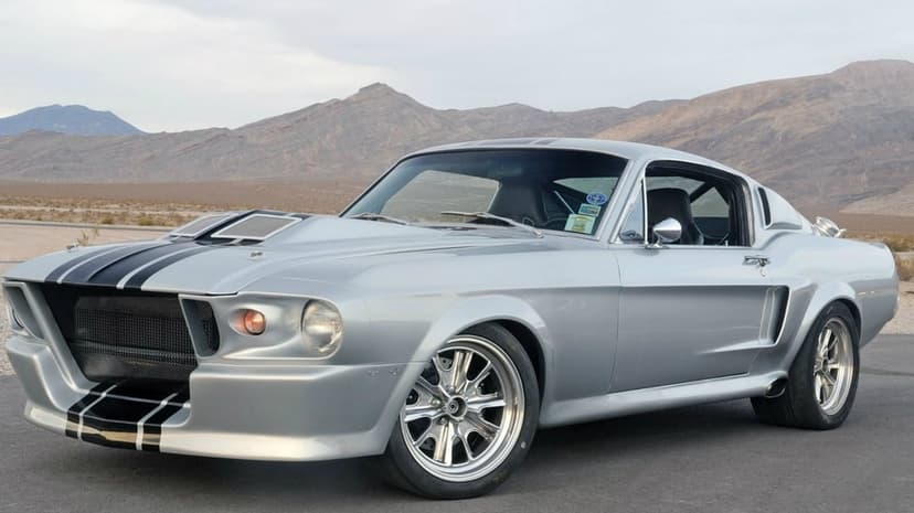 Silver classic Mustang muscle car with black stripes on a desert road with mountains in the background.
