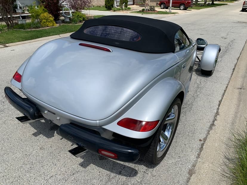Rear view of a silver Plymouth Prowler with a black convertible top and chrome wheels on a sunny asphalt street.