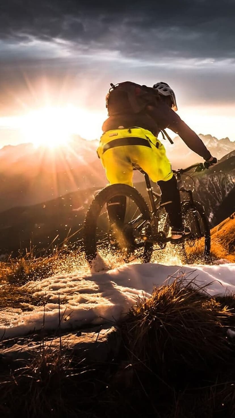 Mountain biker in yellow shorts riding through snow on a mountain ridge with sun rays piercing through clouds.