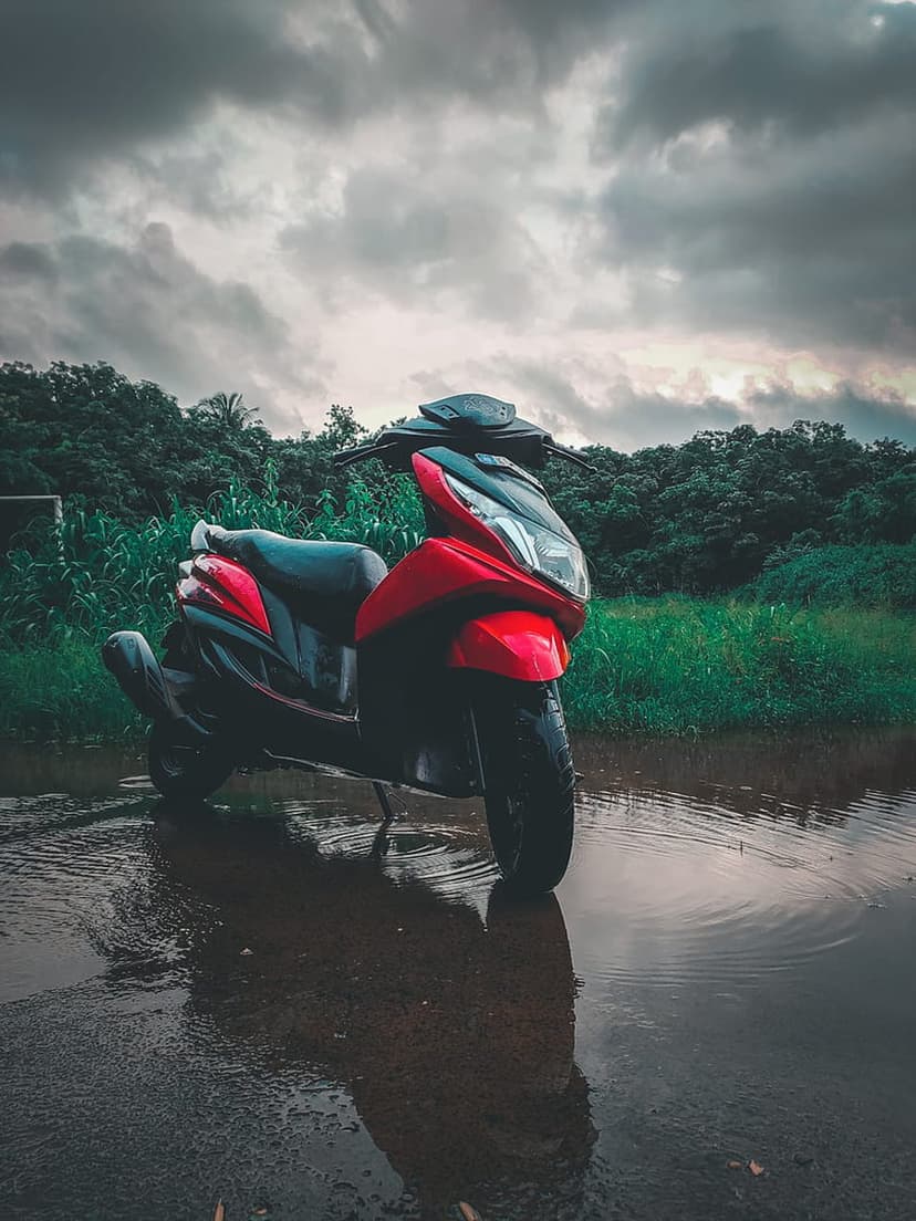 Red scooter parked in a shallow puddle with a clear reflection against a backdrop of green trees and grey clouds.