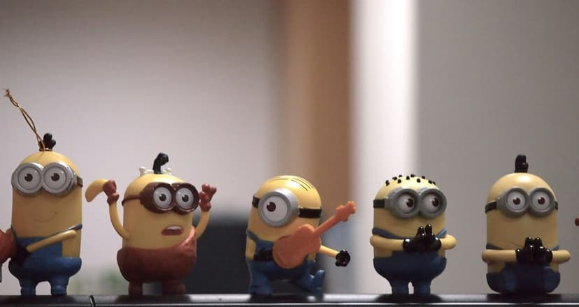 Five Minion toys with yellow skin and blue overalls pose on a shelf, including one character with a guitar.