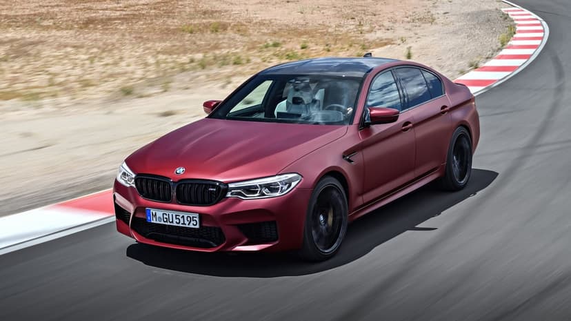 Matte red BMW M5 sports sedan driving at high speed on a curved racetrack with red and white curbs and motion blur.