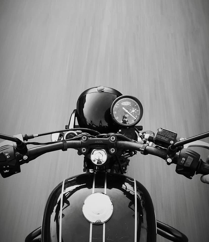 Chrome motorcycle handlebars and a classic circular speedometer are seen from a rider's perspective in grayscale.