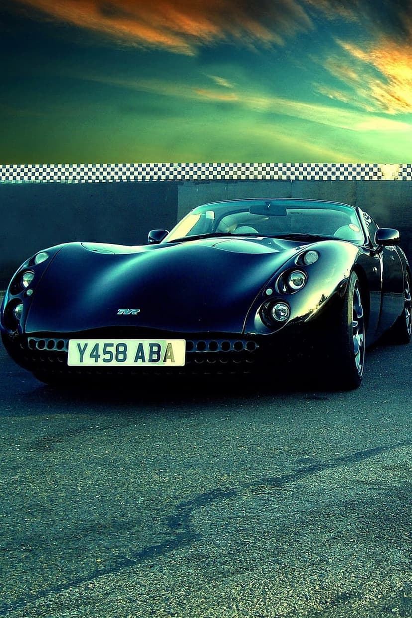 Dark TVR sports car parked on an asphalt race track under a dramatic teal and orange sunset sky at twilight.