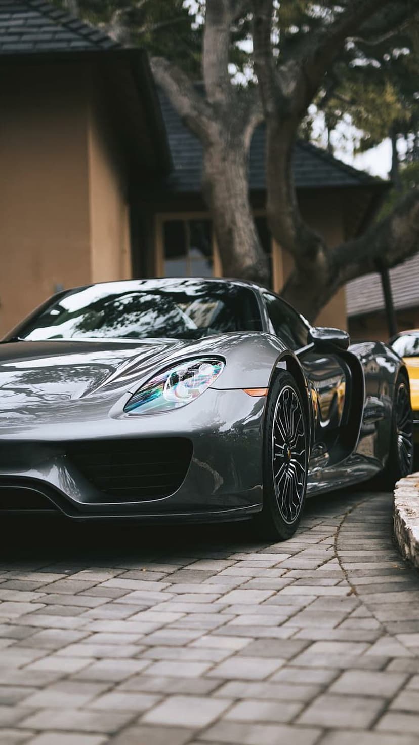 Porsche 918 Spyder in dark grey sits on a cobblestone path under a leafy tree with sharp headlights visible.