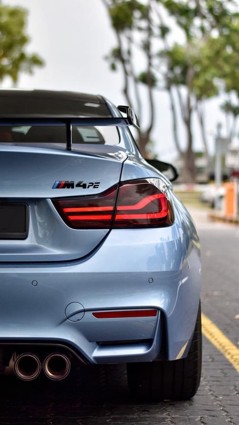 Blue BMW M4 Coupe rear view showing M4PE badge, LED taillights, and dual exhaust pipes against a blurred forest.