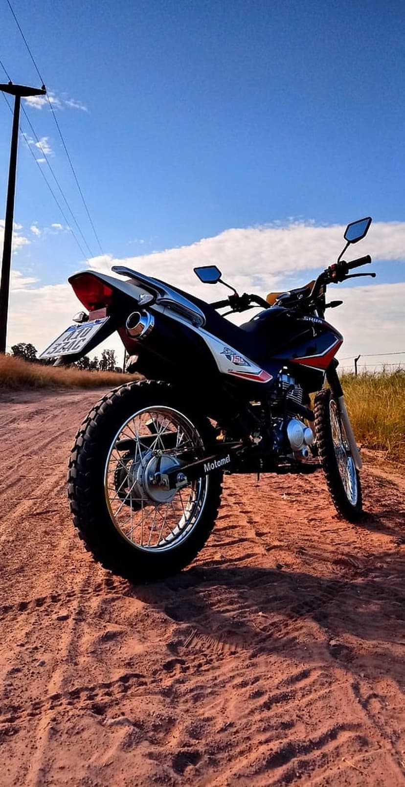 Dirt bike rear wheel and exhaust on a reddish dusty road under a clear blue sky with late afternoon shadows.