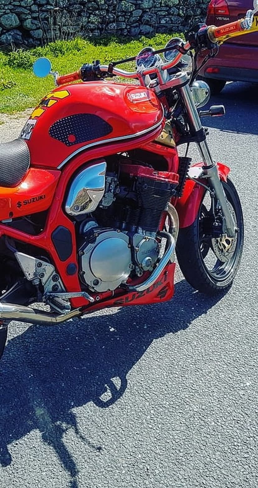 Suzuki motorcycle in red features a detailed engine and handlebars while parked on a sunlit asphalt road outdoors.