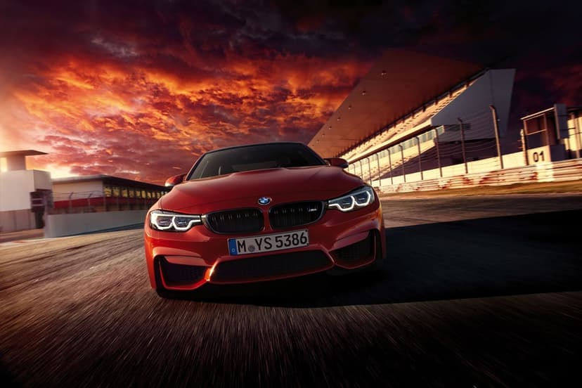 Red BMW M4 racing on a track under a fiery orange sunset with motion blur on the asphalt and glowing headlights.
