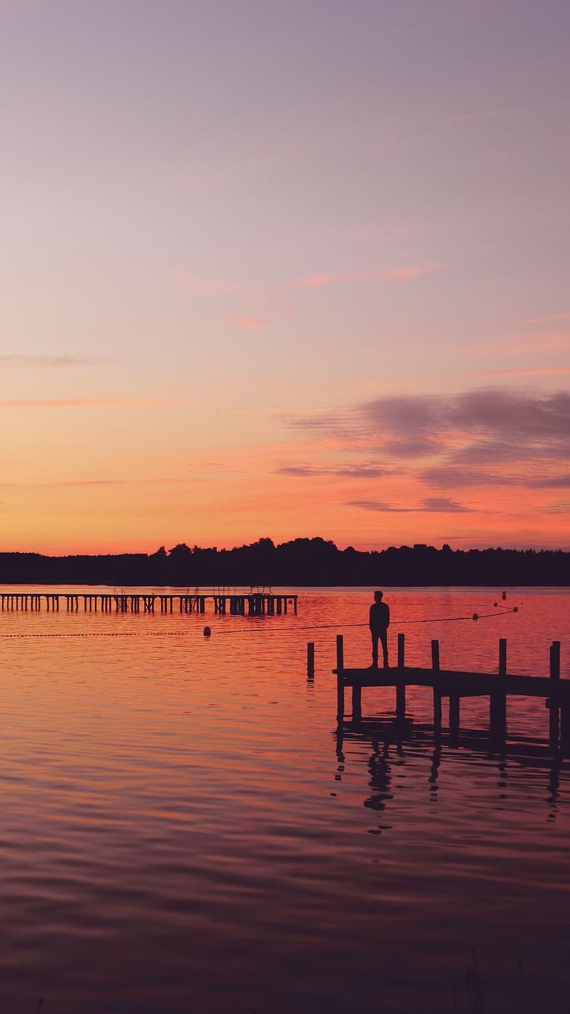 Lone person silhouetted on a wooden pier against a vivid orange and purple sunset reflecting in calm lake water.