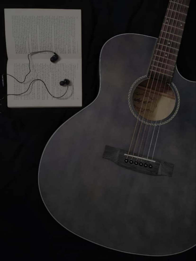 Acoustic guitar sits beside an open book with earbuds on a black surface in a vertical overhead view.