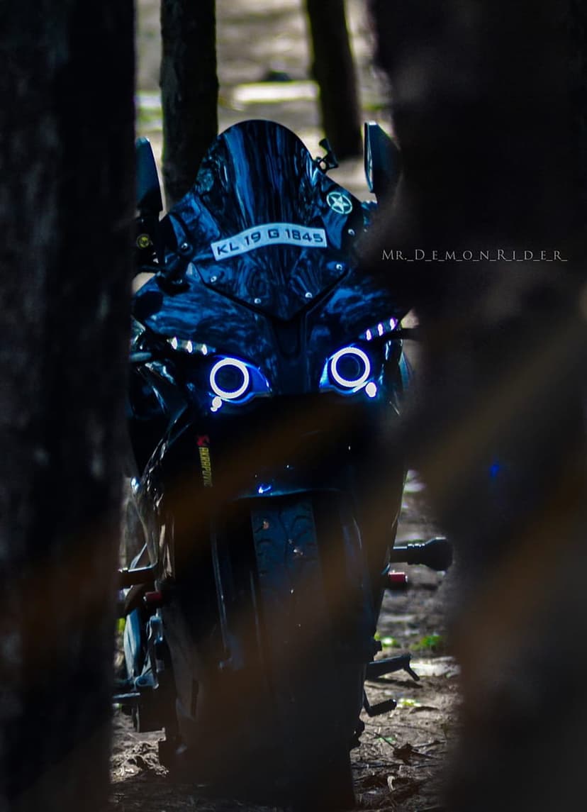 Black motorcycle with glowing blue circular headlights parked in a dark forest at night with visible license plate.