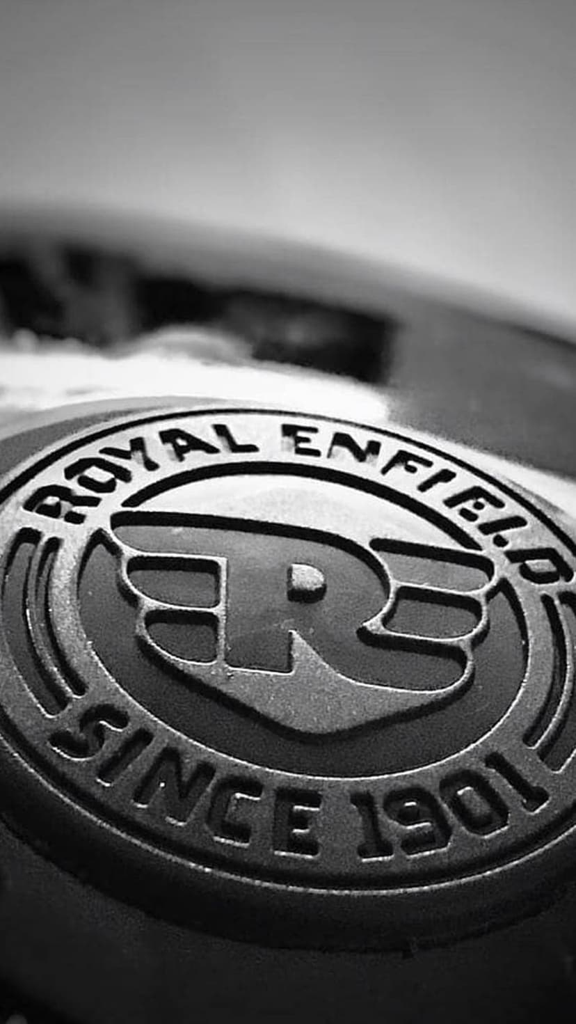 Royal Enfield logo 'R' emblem with circular text on a metallic surface in a black and white close-up shot.