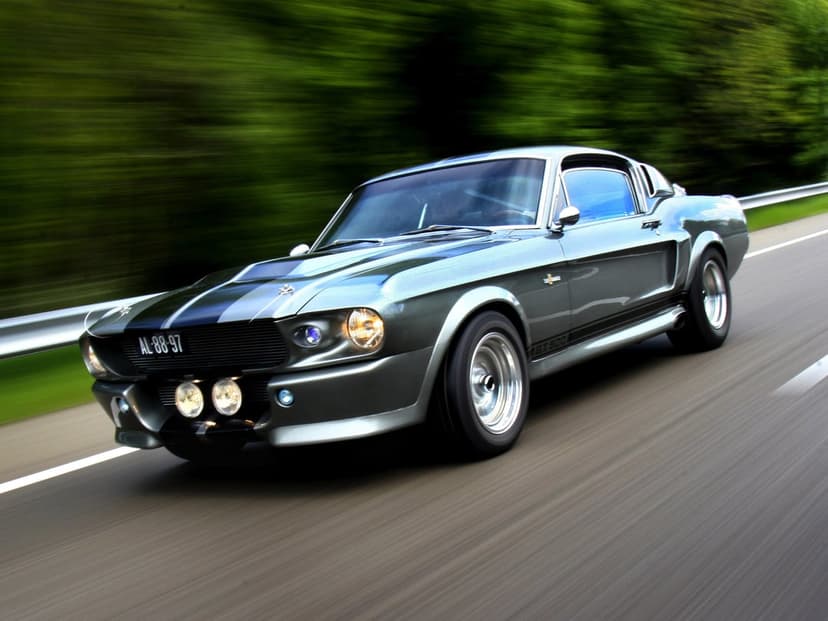 Silver Shelby GT500 Mustang speeding on a highway with motion blur against a lush green forest background.