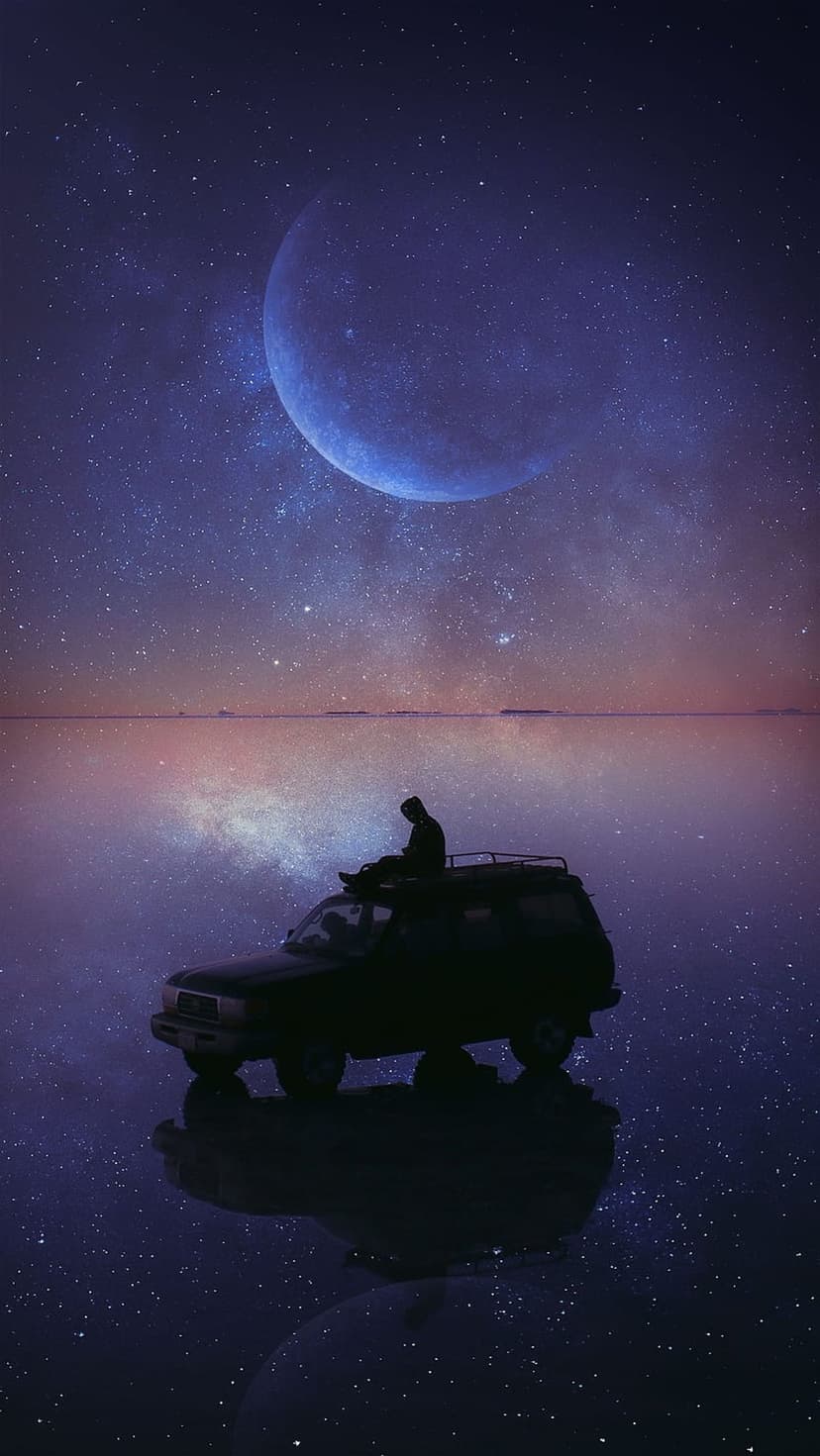 Silhouetted person on an SUV under a giant moon and starry sky reflecting on a perfectly still water surface.