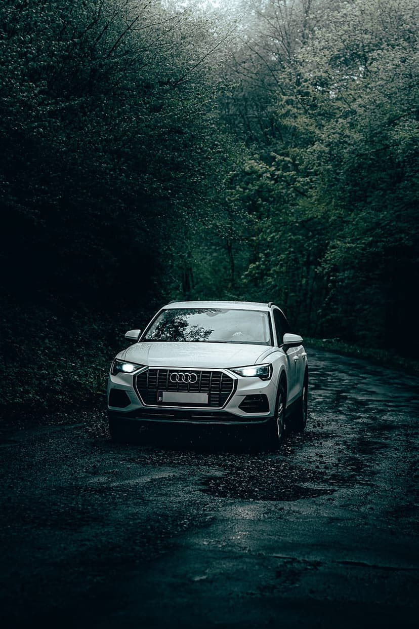 White Audi SUV parked on a dark wet forest road surrounded by dense green pine trees and moody shadows.