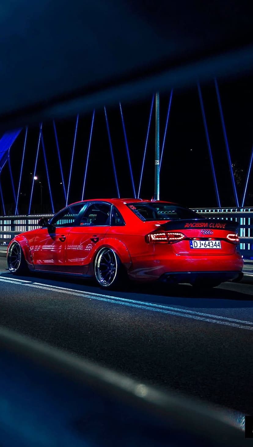 Red Audi S4 sedan with widebody fenders and rear spoiler driving on a bridge at night with blue lights.