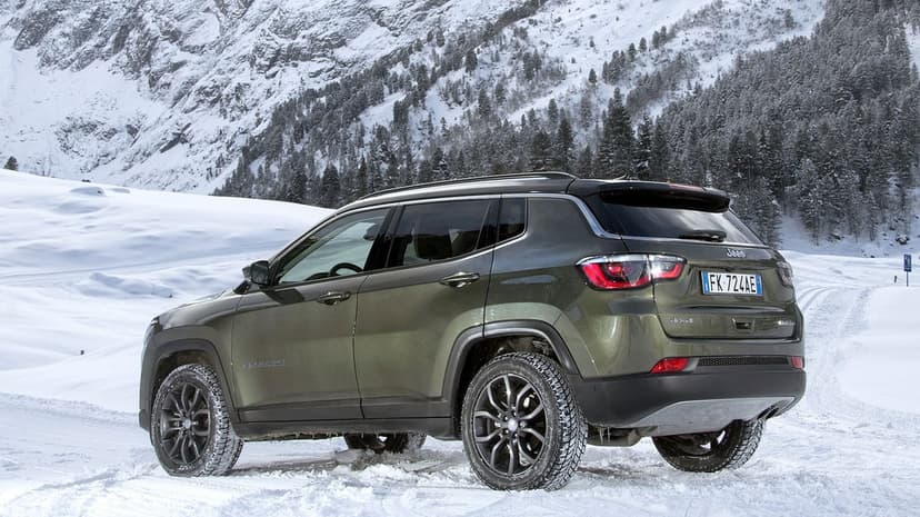 Dark green Jeep Compass parked in deep white snow with towering mountains and pine trees behind it.