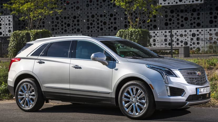 Silver Cadillac XT5 SUV parked on a road with a perforated metal building and green trees in the background.