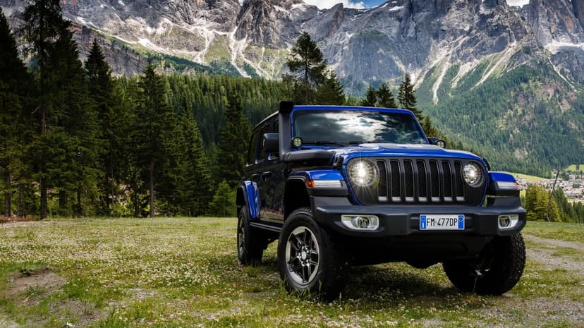 Blue Jeep Wrangler in a grassy field with a backdrop of pine forests and massive rocky mountain peaks.