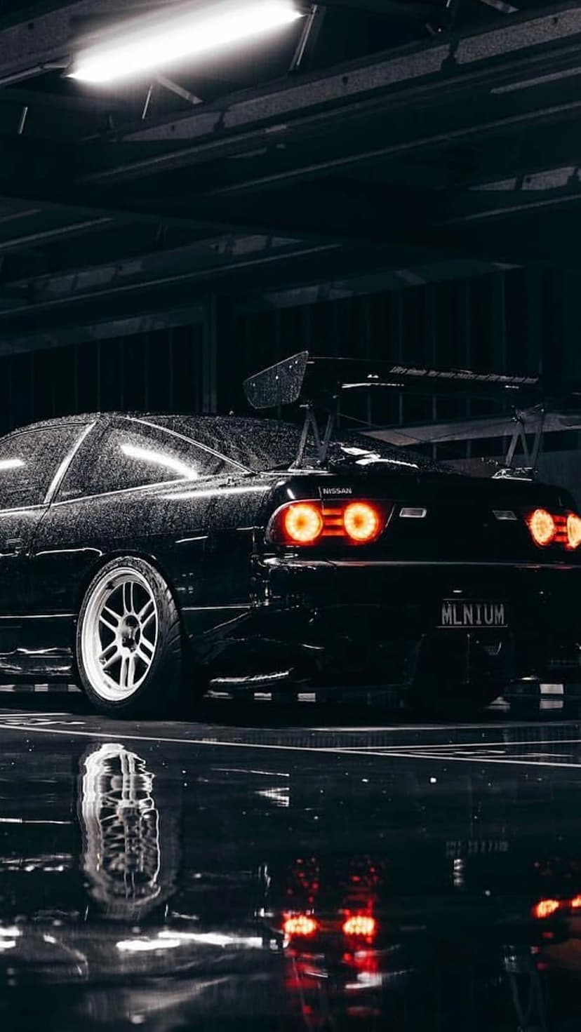 Nissan Skyline GT-R with a rear spoiler sits on wet pavement under night rain with glowing red taillights.