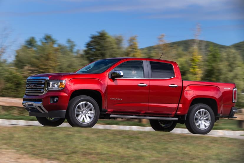 Side profile of a red GMC Canyon pickup truck driving past blurred green trees under a bright blue sky.