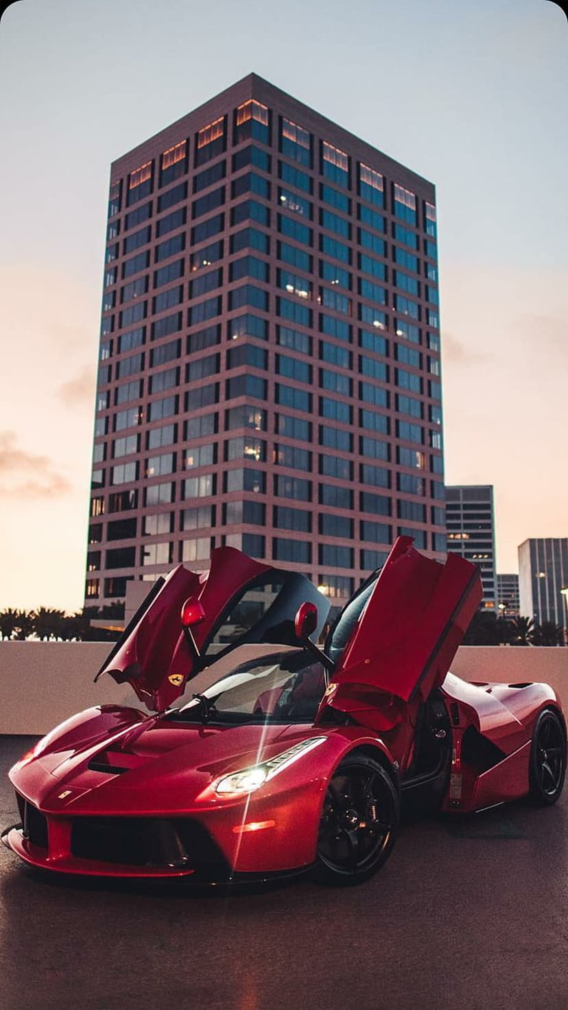 Red Ferrari supercar with vertical doors open parked in front of a glass skyscraper under a twilight sky.