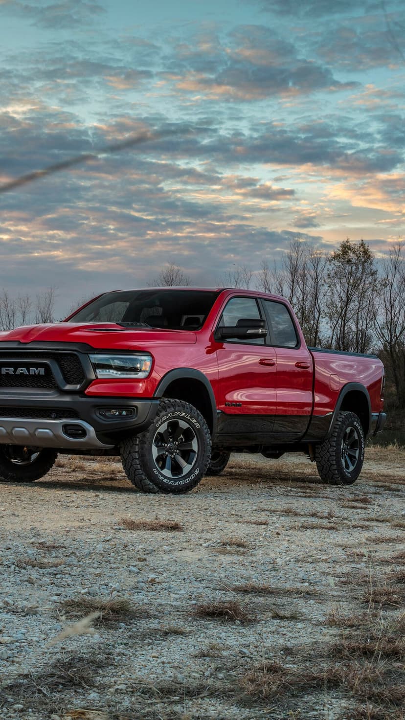 Red Ram 1500 Rebel truck parked on gravel under a vibrant cloudy sunset sky with bare trees in the background.