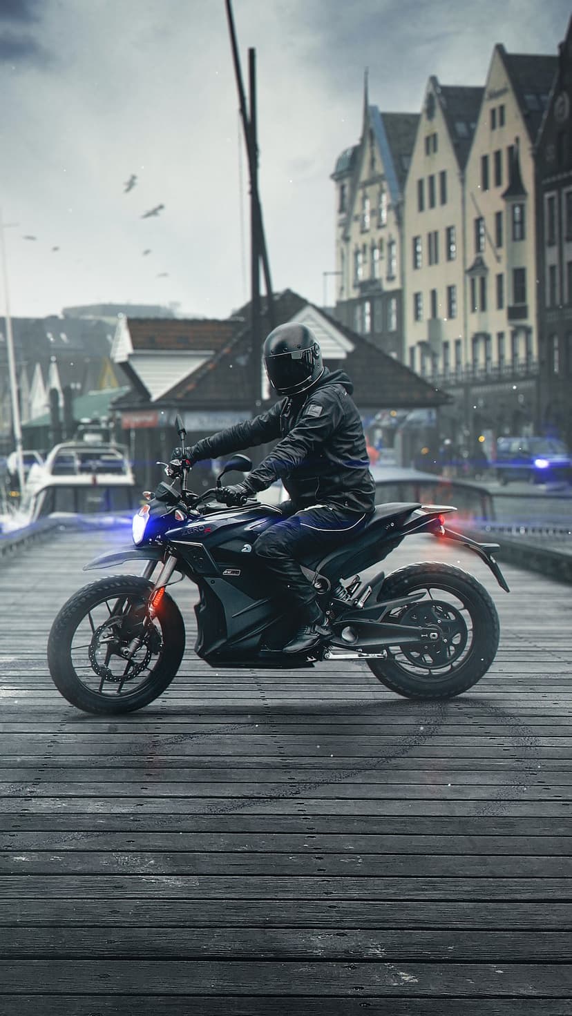 Motorcyclist on an electric bike with a blue headlight rides across a wooden pier near a cloudy urban waterfront.