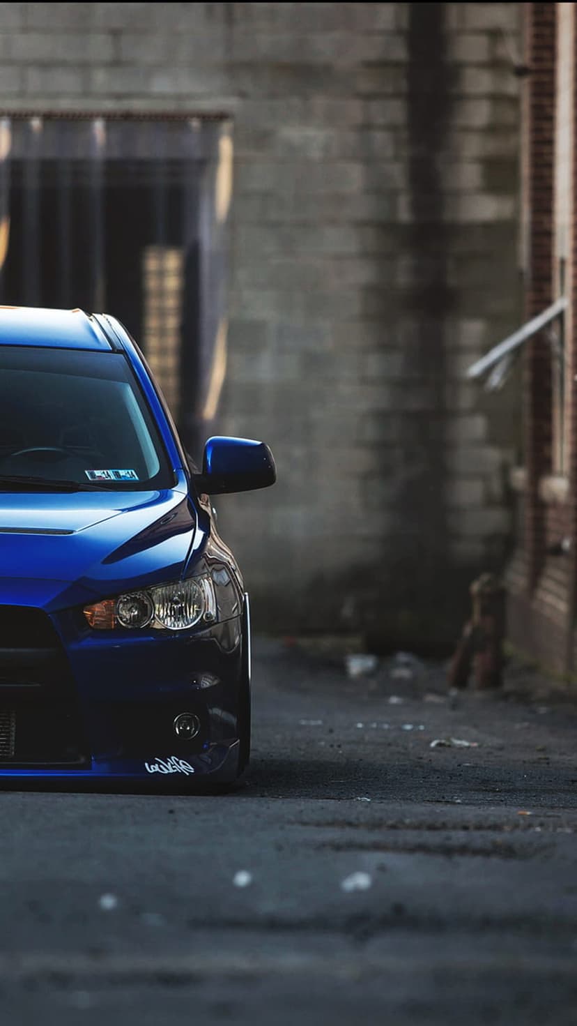 Blue Mitsubishi Lancer Evolution front bumper parked in a dark brick alley with low-angle lighting and soft bokeh.