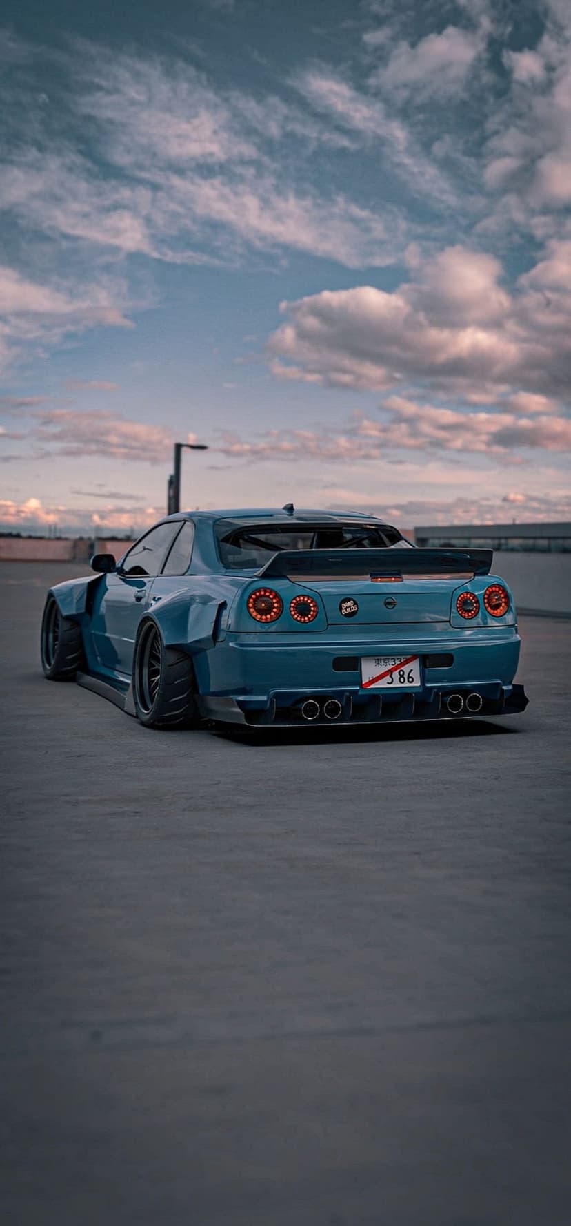 Nissan Skyline R34 GT-R rear view showing widebody fenders and glowing circular taillights on a concrete surface.