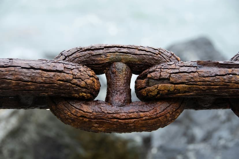 Thick rusted iron chain links with a textured orange patina over a bokeh background of grey rocks and water.