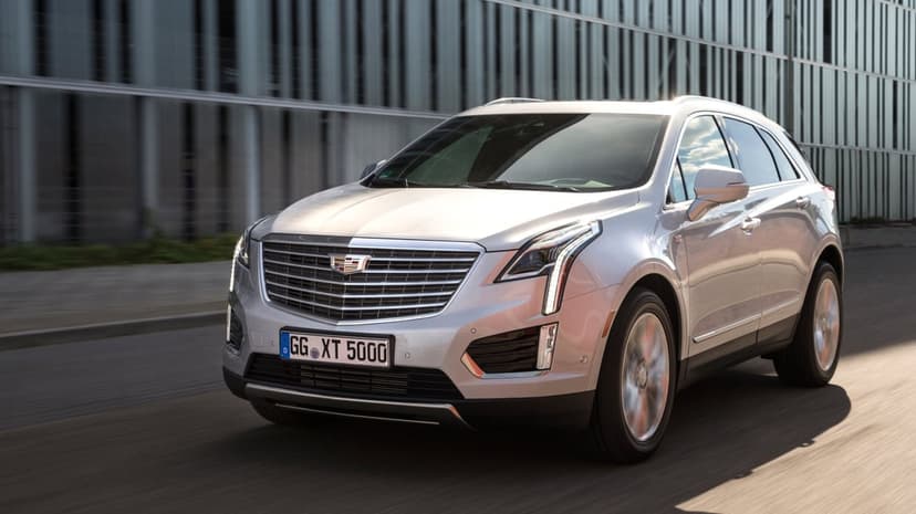 Silver Cadillac XT5 SUV driving on a city street in front of a modern building with vertical metal slats.