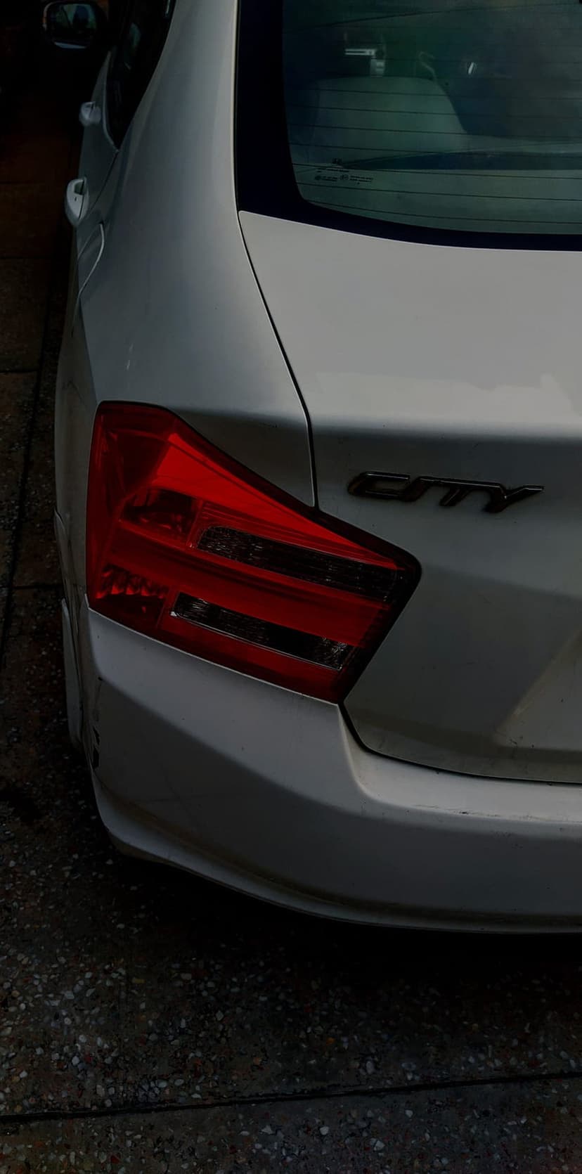 Honda City red tail light and black emblem on a white rear car panel over grainy asphalt ground textures.