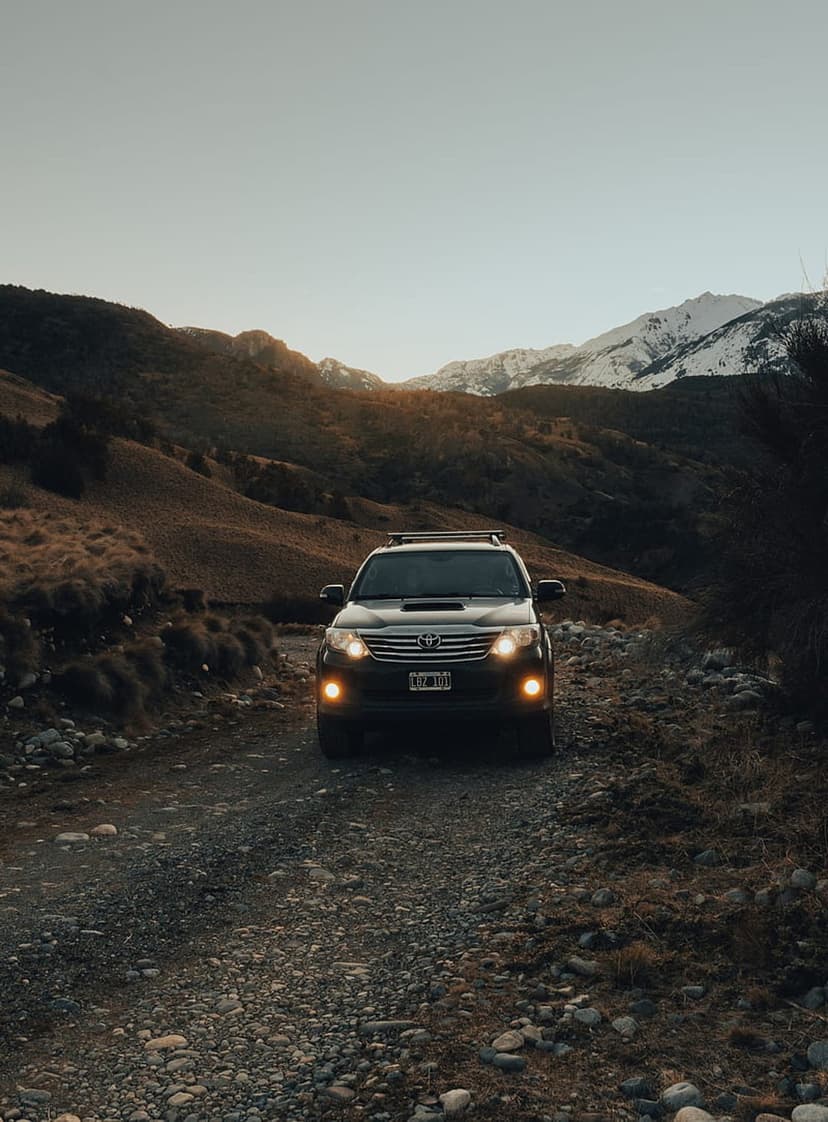 Dark SUV parked on a gravel mountain road with snow-covered peaks under a dusky purple and orange sunset sky.