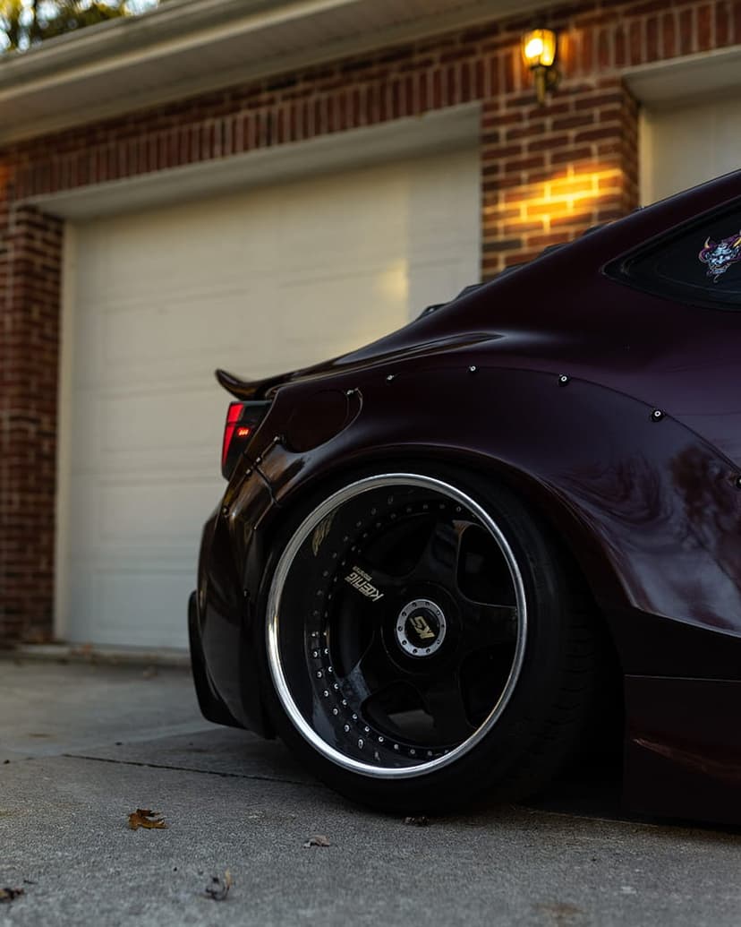 Purple widebody sports car with black deep-dish wheels parked in front of a brick building with a white door.