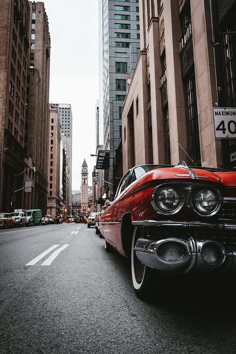 Red Cadillac with chrome details parked on a city street next to vintage cars and a speed limit sign.