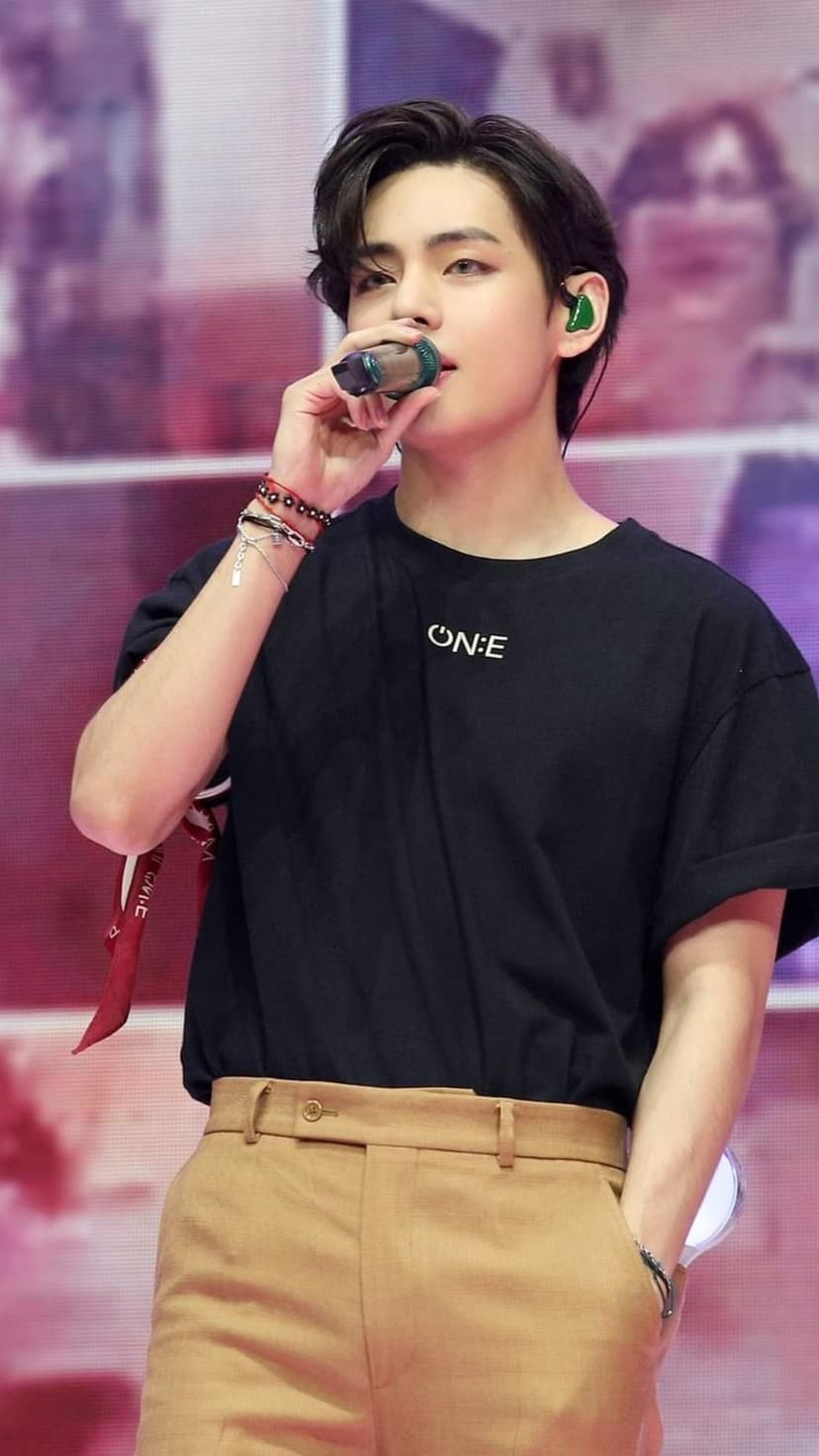 Kim Taehyung sings into a microphone while wearing a black t-shirt and green in-ear monitors on a concert stage.