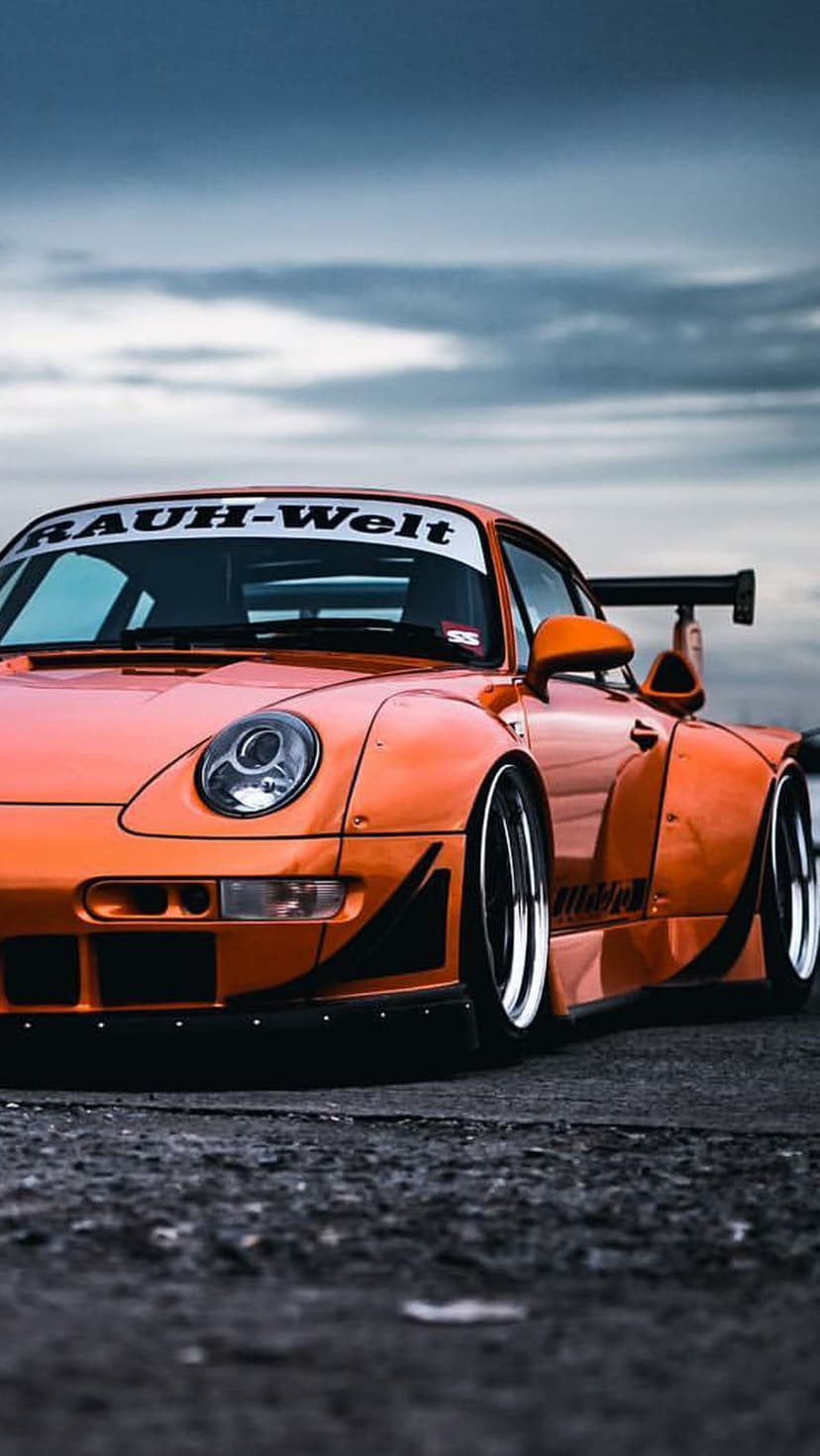 Orange Porsche 911 RWB with wide fenders and a large rear wing parked on asphalt under a cloudy sky.