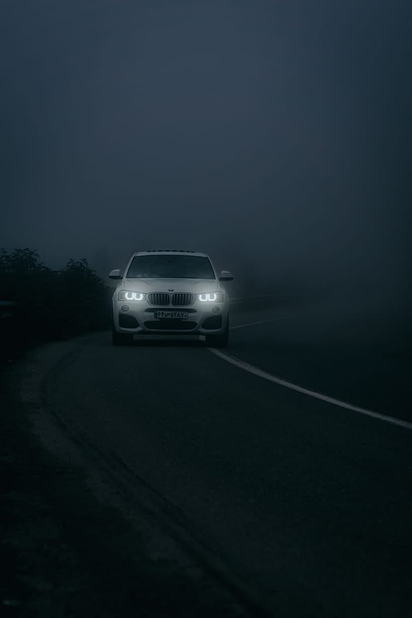 White BMW SUV with glowing headlights driving through heavy mist on a dark asphalt road during the night.