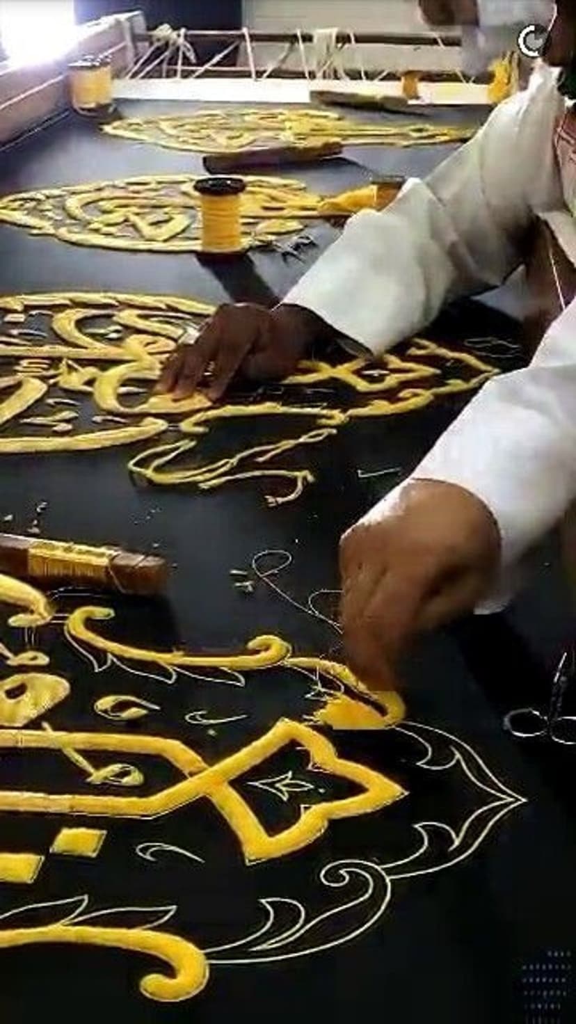 Golden Arabic calligraphy embroidery on black fabric with thread spools and an artisan's hands working.