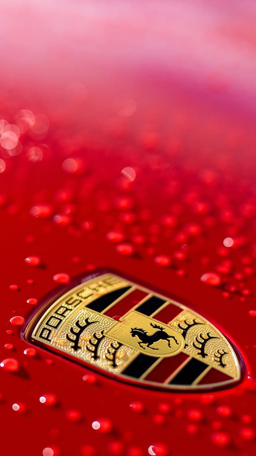 Porsche emblem with gold and black details covered in water droplets against a glossy red metal car surface.