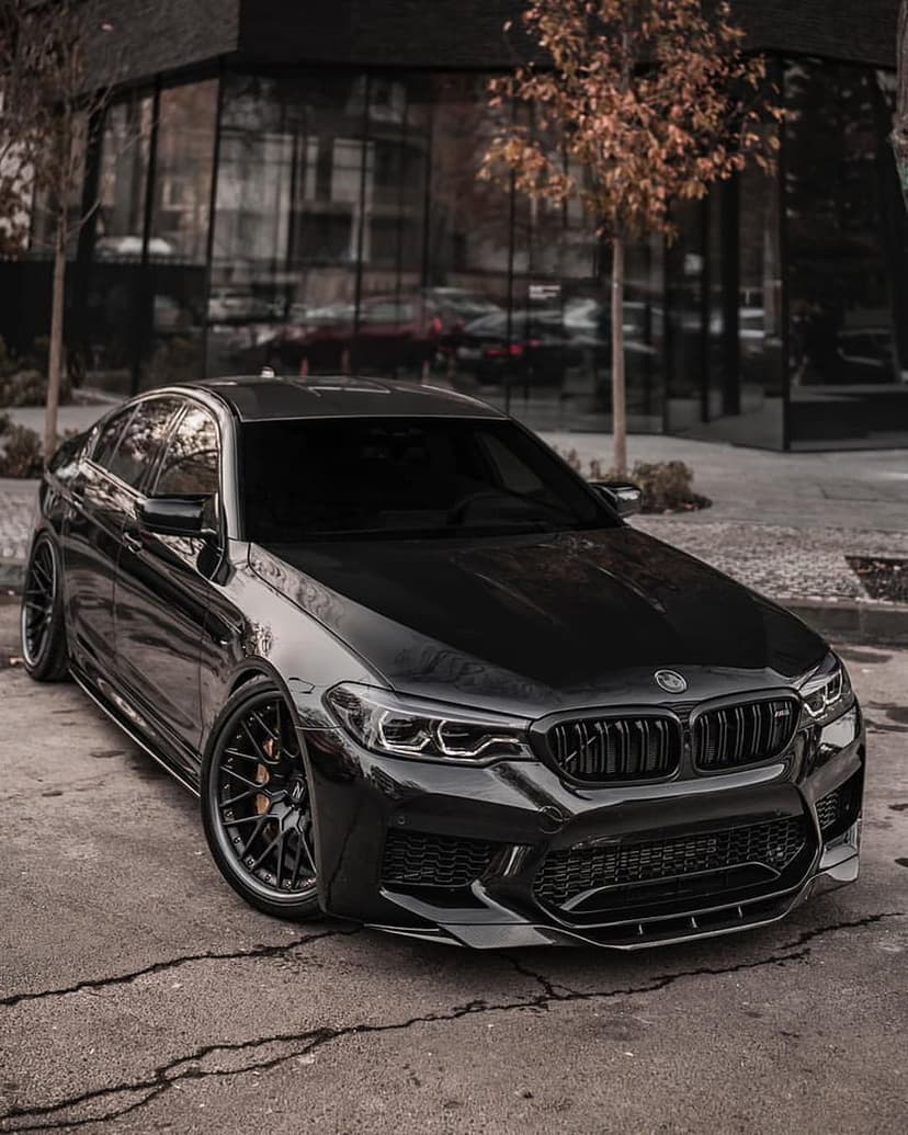 BMW M5 black sedan parked on cracked asphalt with autumn trees and a glass building in the urban background.