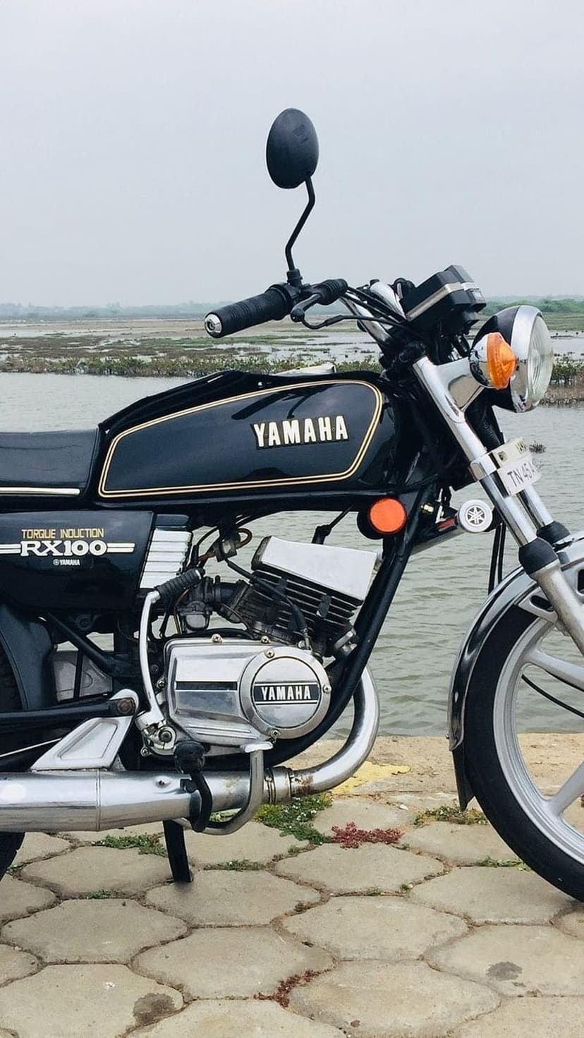 Black Yamaha RX100 motorcycle with chrome accents parked on a paved path next to a serene lake and green trees.