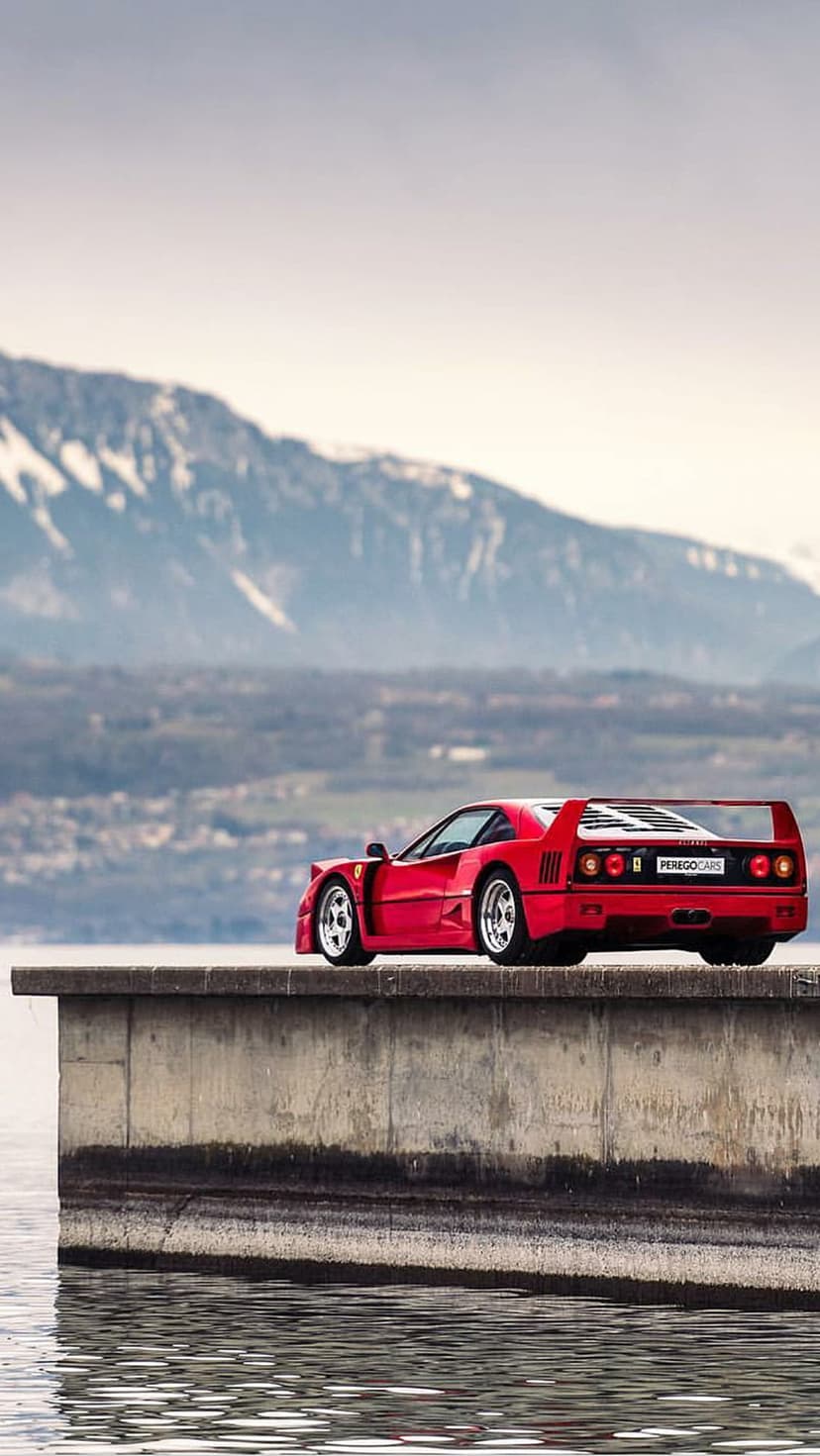 Red Ferrari F40 sports car parked on a concrete ledge by calm water with snow-capped mountains in the background.
