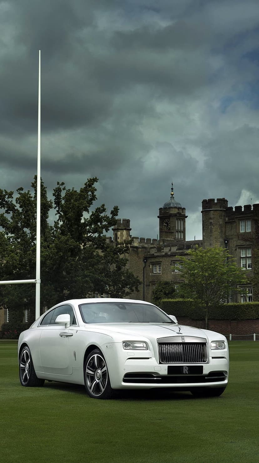 White Rolls-Royce Wraith parked on green grass with a stone clock tower castle under a moody, overcast sky.